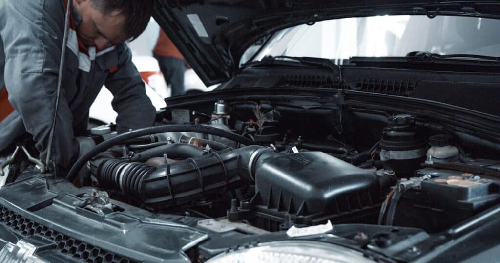 A mechanic in uniform works under the raised hood of a car, inspecting or repairing the engine components.