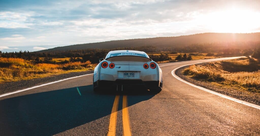 A white sports car drives down a winding rural road at sunset with fields and hills in the background.