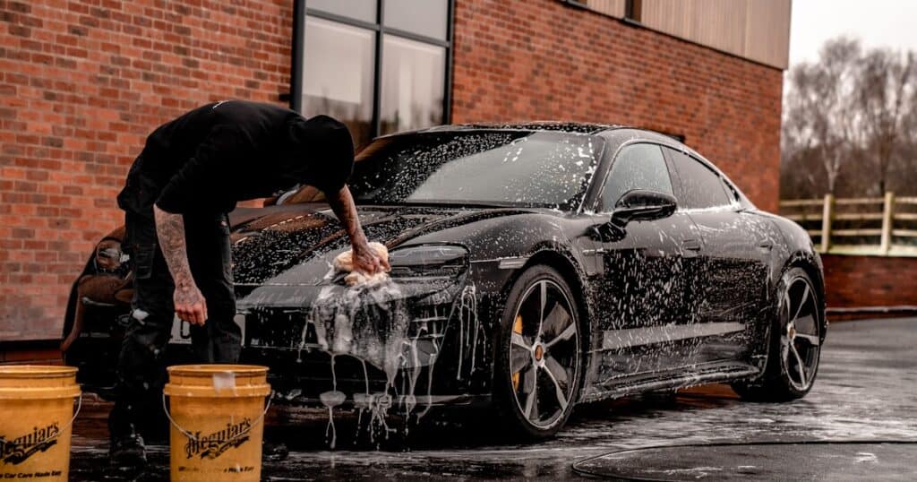 A person washes a black sports car with soap and water using a sponge, next to two yellow buckets, in an outdoor area by a brick building.