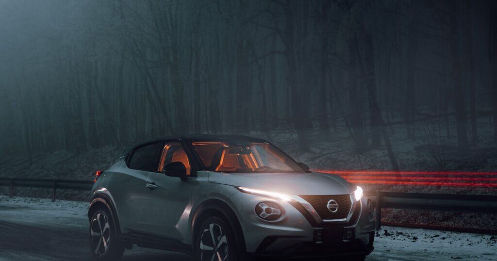 A silver Nissan SUV with headlights on is parked on a snowy road at night, with a dark, foggy forest in the background and red light trails passing by.