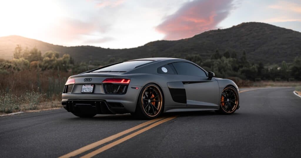 A matte gray Audi R8 sports car is parked on a two-lane road with mountains and trees in the background at sunset.